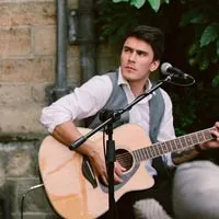 Young man playing acoustic guitar and singing into microphone outdoors against a brick wall and green foliage.