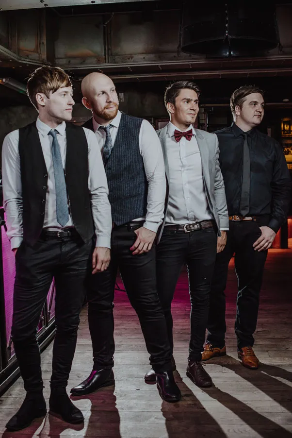 Four men dressed formally in shirts, ties, and vests or jackets standing indoors on a wooden floor under industrial lighting.
