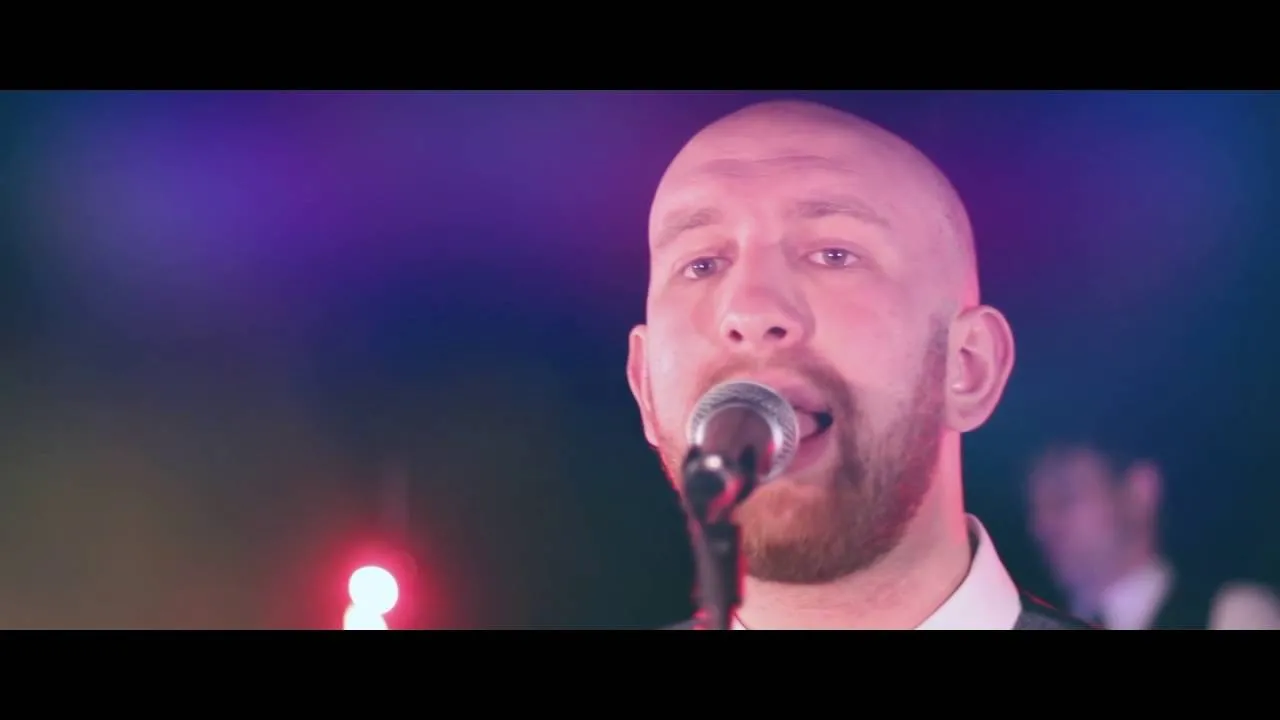 Close-up of a bald man with a red beard singing into a microphone under purple and pink stage lighting.