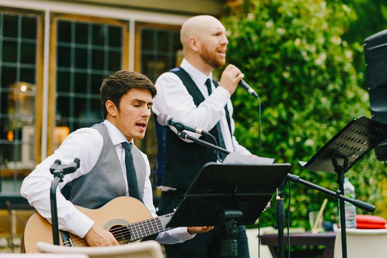 Two men in white shirts and vests performing music, one playing acoustic guitar and singing into a microphone, the other singing with a microphone.