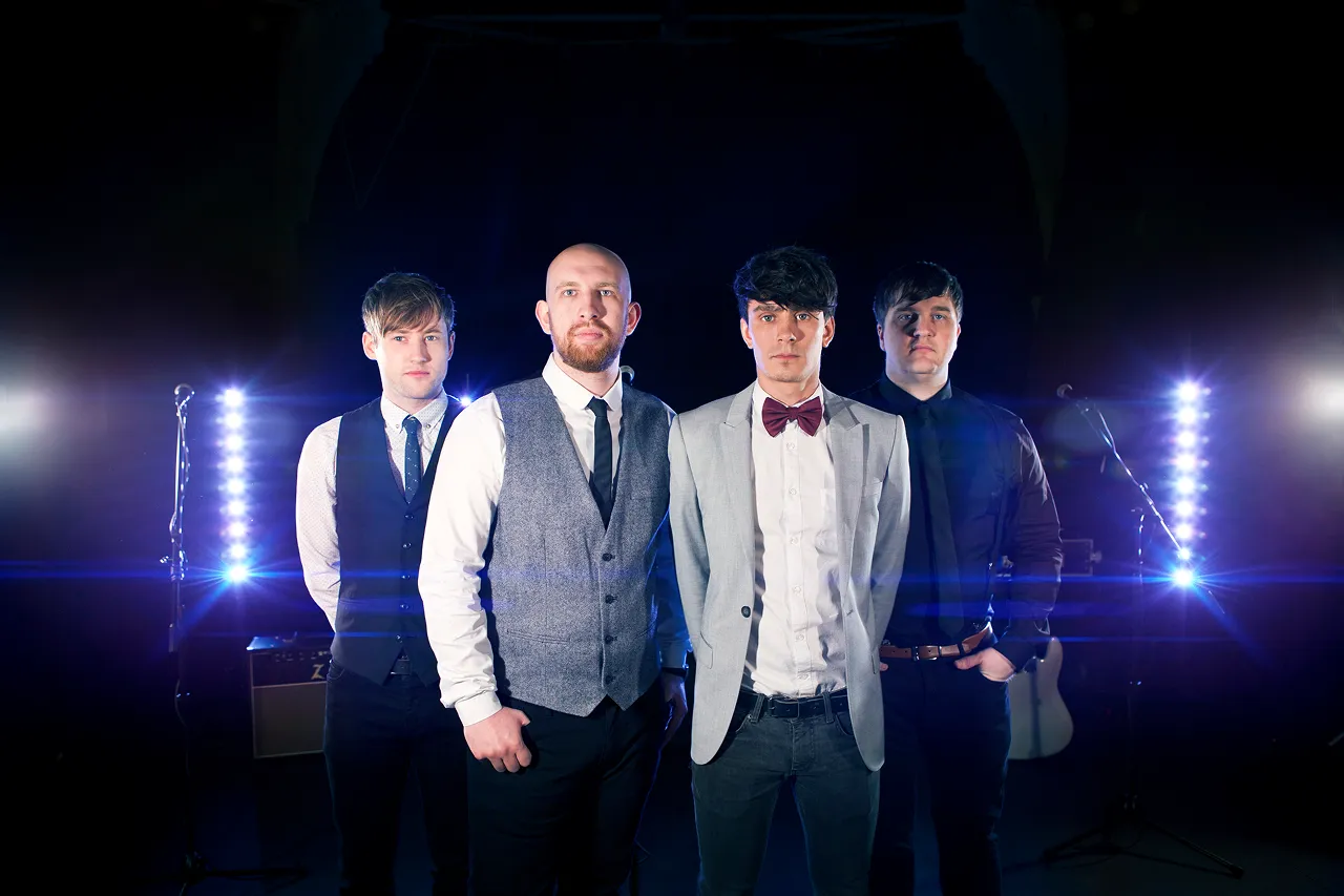 Four men dressed in formal attire standing in a line with stage lights and microphones behind them.